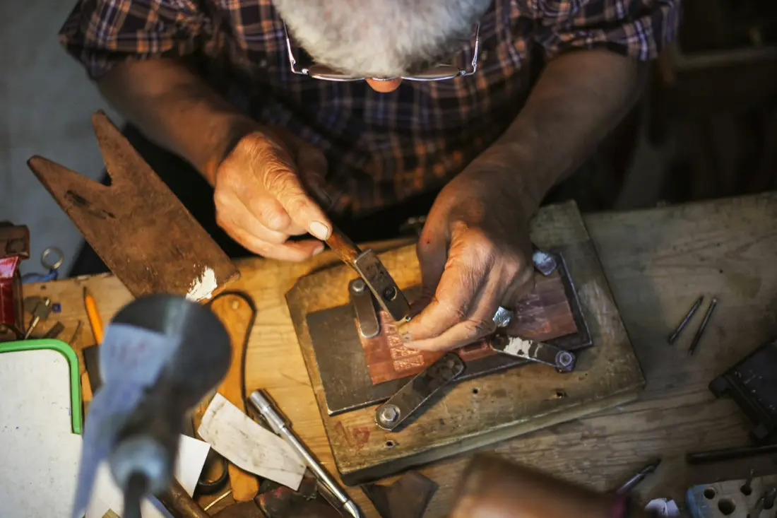 Manos de un orfebre trabajando una pieza de metal sobre el banco del taller, rodeadas de herramientas.
